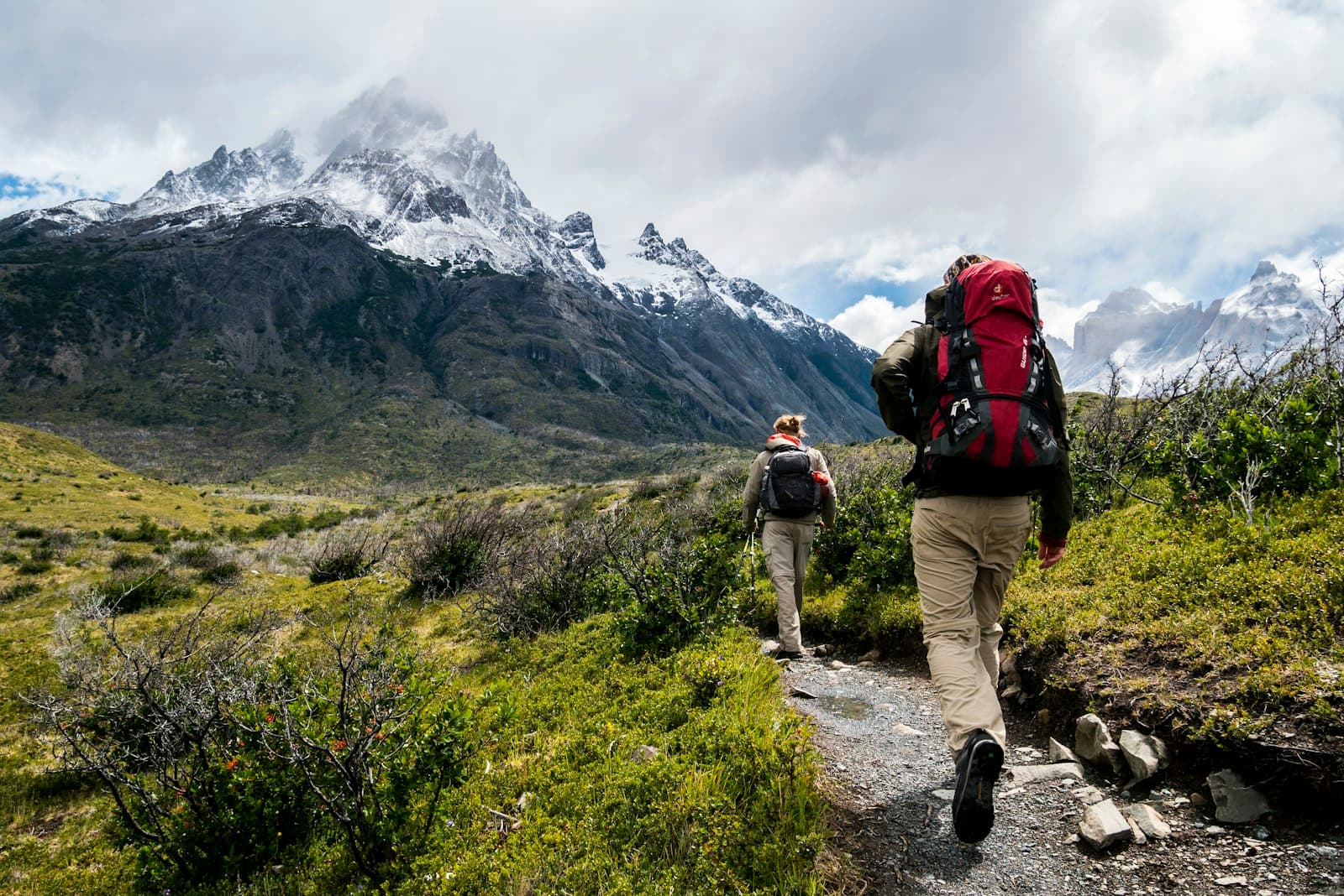 Two hikers with backpacks on a singletrack trail below jagged snow-capped peaks